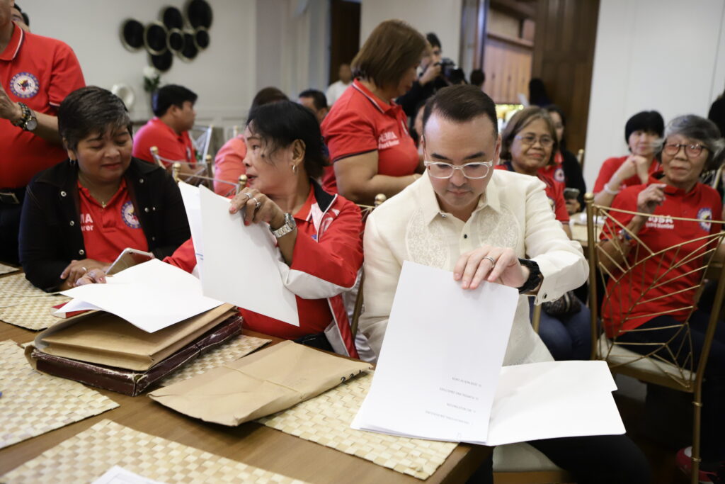 Alan Peter Cayetano reviews documents with a group in red shirts, focusing on details, collaborating, and engaging in policy review or planning.
