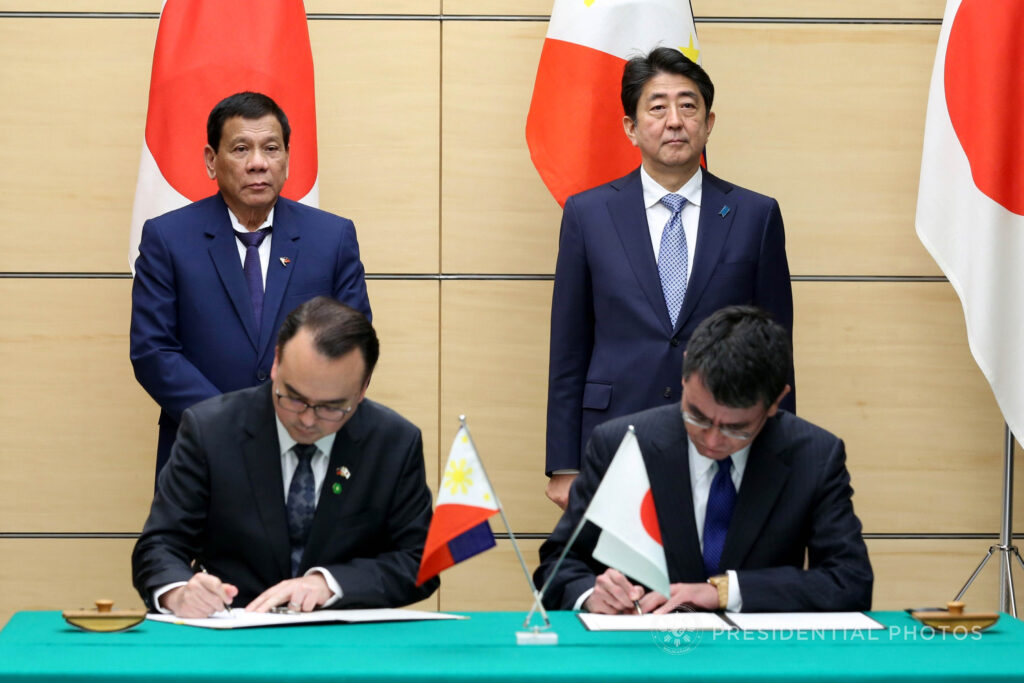 Alan Peter Cayetano signs an agreement alongside a Japanese representative, with Duterte and Abe observing the ceremony.