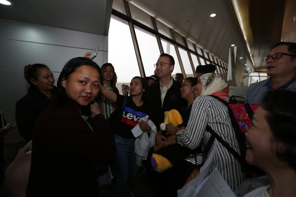 Alan Peter Cayetano poses for photos with a group of Filipinos, possibly OFWs, at an airport, showing camaraderie and support.